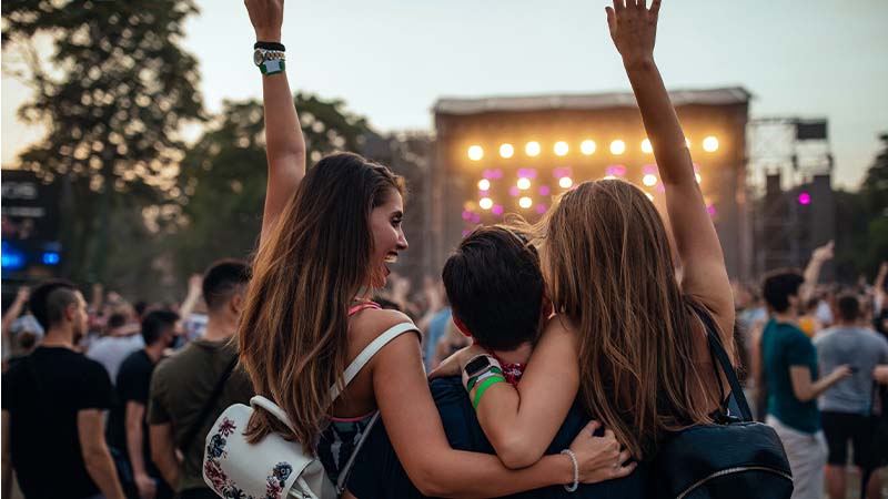 Multidão num concerto ao ar livre; várias pessoas de costas com braços no ar, palco iluminado e público ao fundo.